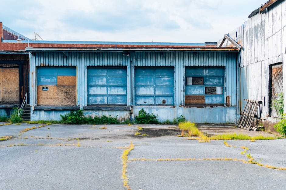 A decaying industrial warehouse with boarded-up windows and overgrown pavement.